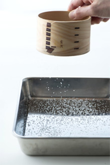 写真：テーブル上の銀色のバットに、20cmぐらい上から小さいふるいに入れた塩を振りかけている